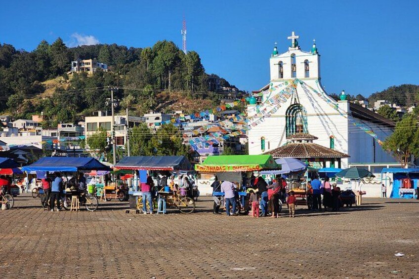 A bustling outdoor market with vendors, a prominent church, and a hilly backdrop. 5