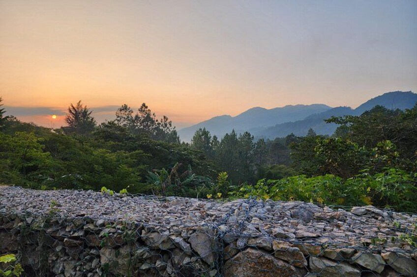 A stone wall, dense green foliage, and distant mountains during sunset. 4