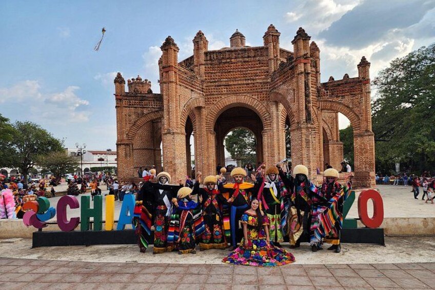 A group of people in colorful traditional attire posing in front of a large brick archway with the letters 'CH' and 'O' visible. 1