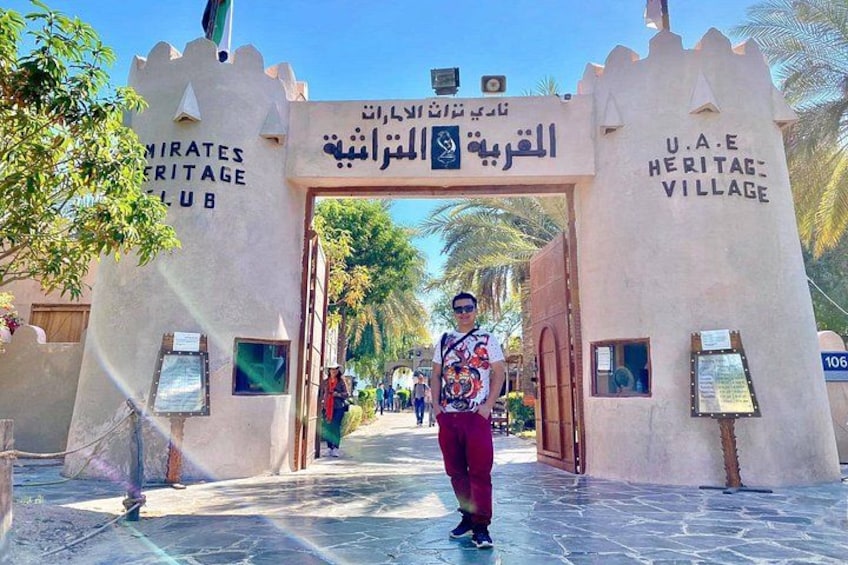 A man standing in front of a gate with the text 'U.A.E. Heritage Village' and 'Emirates Heritage Club' on it. 2