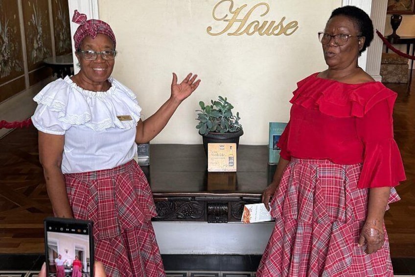 Deux femmes, vêtues de tenues traditionnelles, se tiennent dans une pièce où est affiché un panneau indiquant « Maison ». 2
