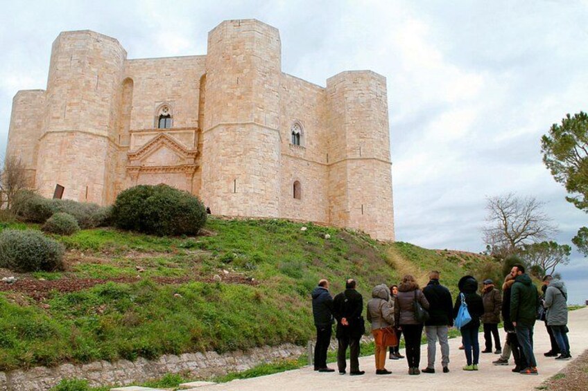 A historic stone castle with battlements, a group of people standing in front, and a grassy hill. 3