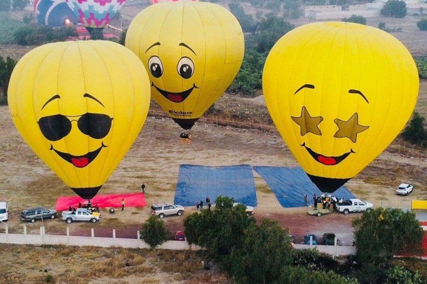 Three hot air balloons with smiling faces, parked on a dirt field with tents and vehicles in the background. 2