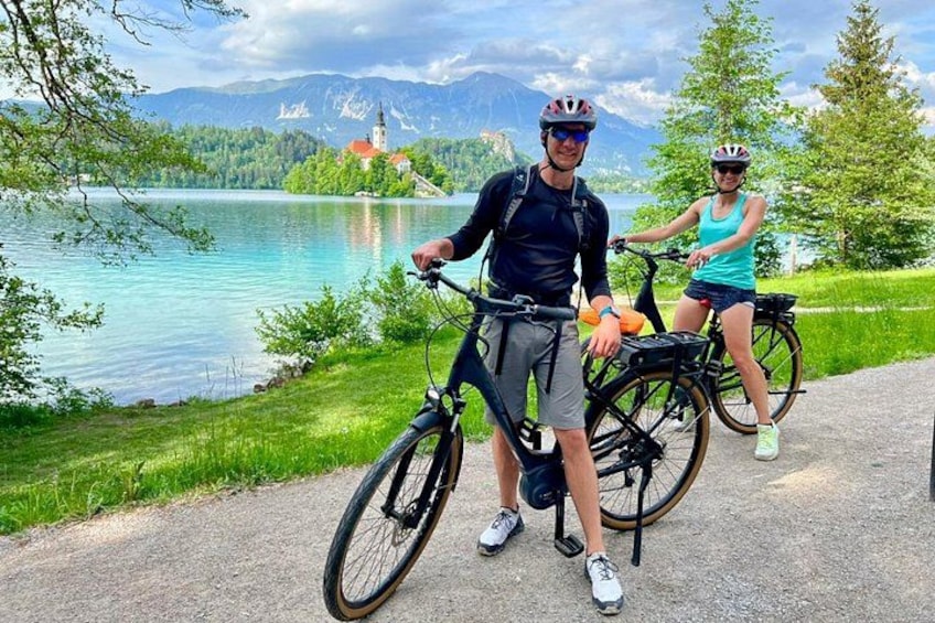Dos personas en bicicleta cerca de un lago, con una iglesia y montañas al fondo. 1