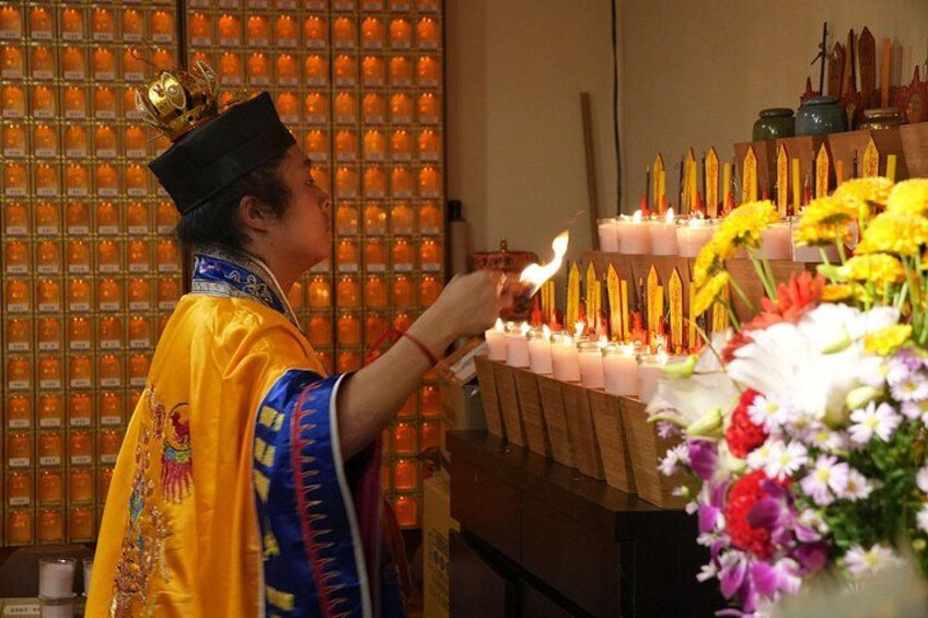A person in traditional attire lighting candles in a room with a wall of incense burners. 2