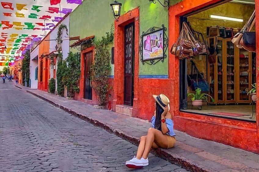 A street with colorful buildings, a person sitting on the sidewalk, and hanging decorations. 3