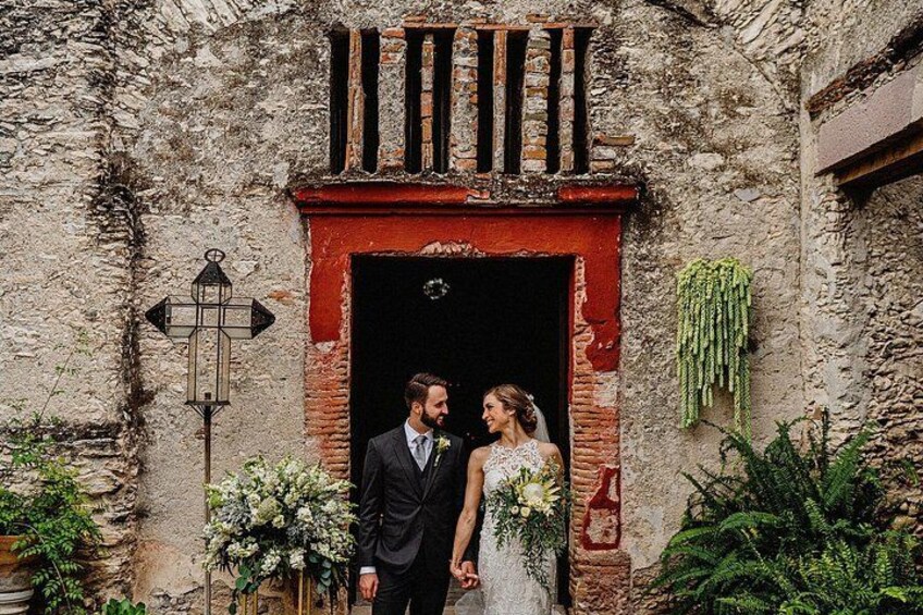 A couple exiting a rustic building with a brick facade and a red archway. 2