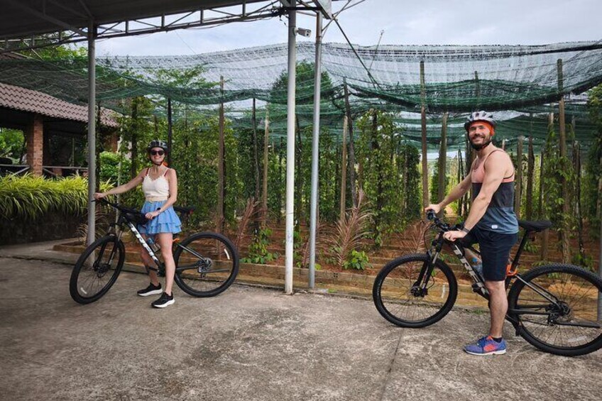 Two people standing next to bicycles in a covered area with plants and a greenhouse structure in the background. 3