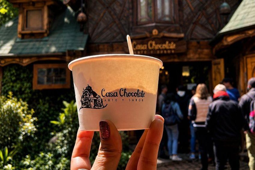 A hand holding a cup with 'Casa Chocolate' text, a building with a sign in the background, and people walking. 5