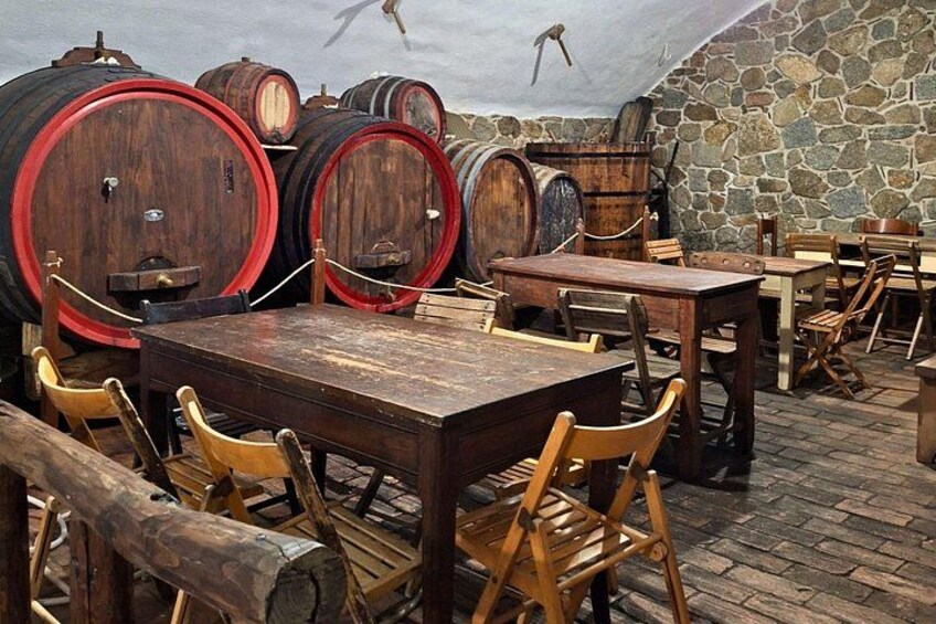 A rustic cellar with wooden barrels, stone walls, and wooden tables and chairs. 3