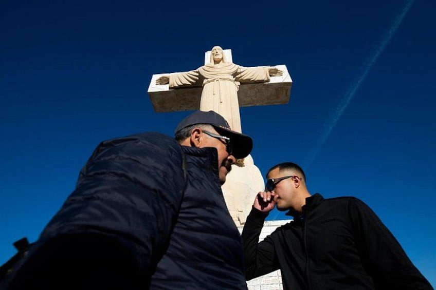 A large stone cross with a figure at the top, two people standing in front of it, and a clear blue sky in the background. 2