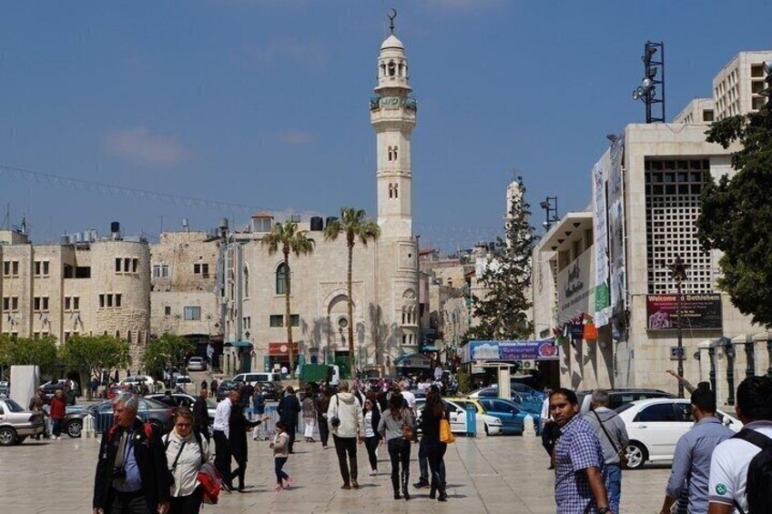 A busy urban square with a prominent mosque featuring a tall minaret, surrounded by modern buildings and pedestrians. 1