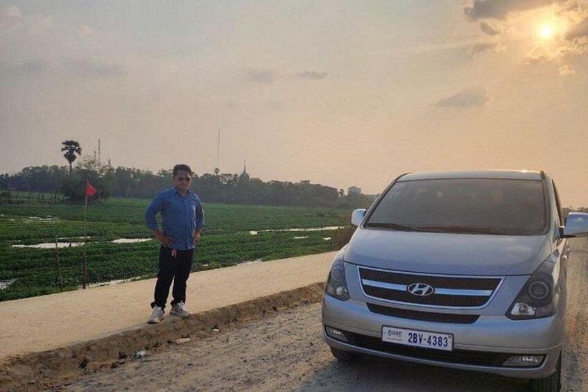 A person standing next to a silver Hyundai car on a rural road with fields on the side. 4