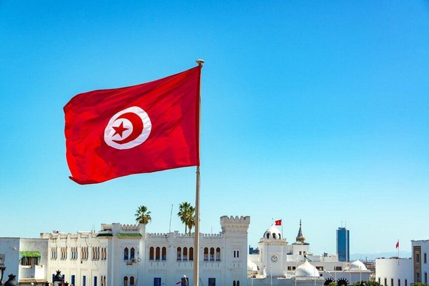 A red flag with a white circle and star, a white building with arches, and a clear blue sky. 1