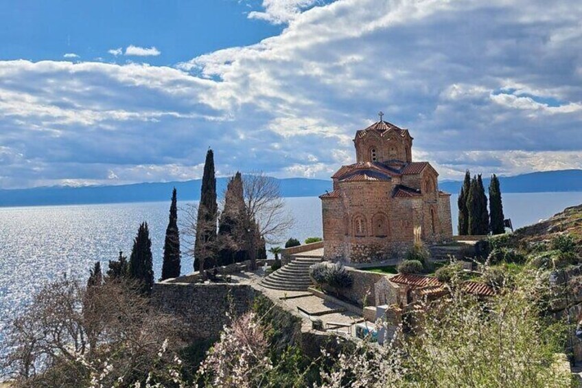 A historic church with a bell tower, surrounded by cypress trees and blooming shrubs, overlooking a body of water. 2