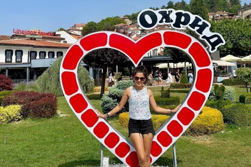 Una persona de pie frente a un letrero con forma de corazón que muestra la palabra "Окрхид" en un parque con follaje y edificios al fondo. 5