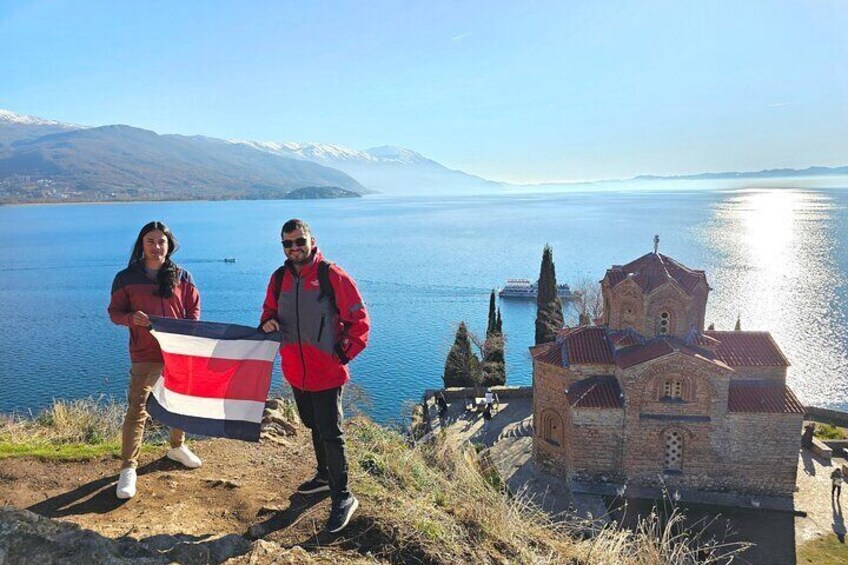 Dos personas sosteniendo una bandera, una iglesia y un lago con montañas al fondo. 2