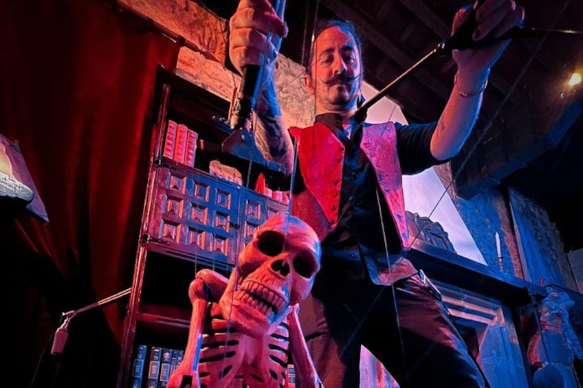A person in a red vest holding a microphone, standing in a room with a skeleton prop and a bookshelf in the background. 1