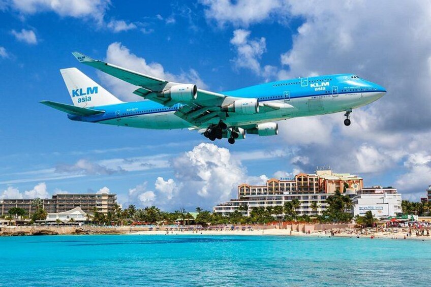 Et KLM Asia flyvemaskine letter over en strand med bygninger og palmer i baggrunden. 1