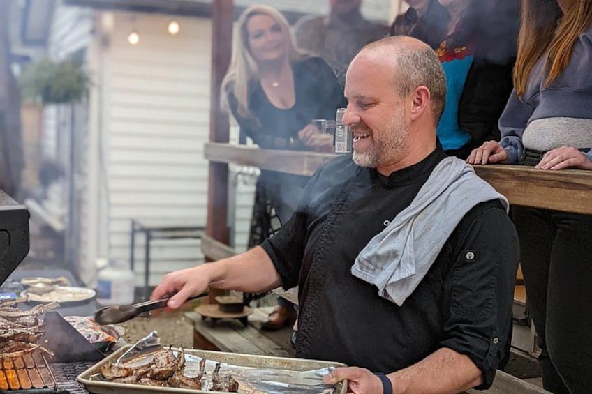 A chef grilling food outdoors with a crowd watching. 2