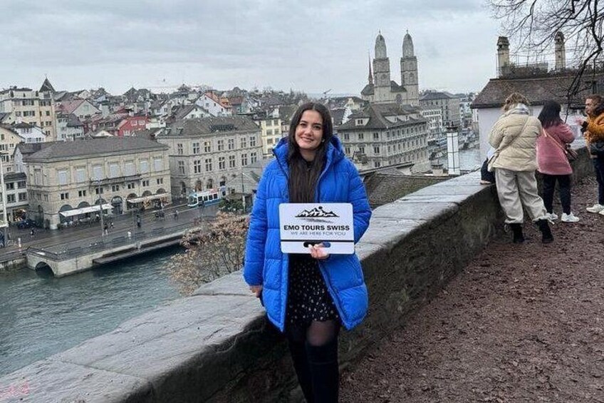 A person holding a sign with 'EMO TOUR SWISS' text, standing on a stone bridge with a cityscape in the background. 1