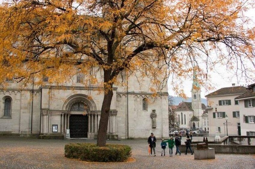 A historic building with a large tree in front, a paved courtyard, and a group of people. 3