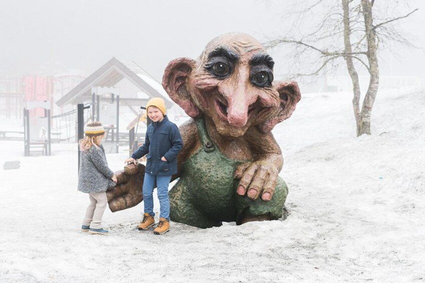 A snowy landscape with a large statue of a creature, two people, and a wooden structure in the background. 2