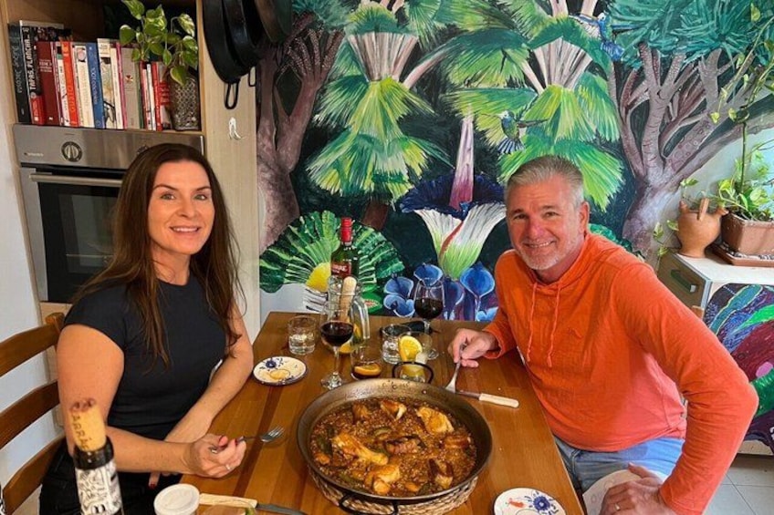 Two people sitting at a table with a large pan of food, a bottle of wine, and a painting of a tropical scene in the background. 3