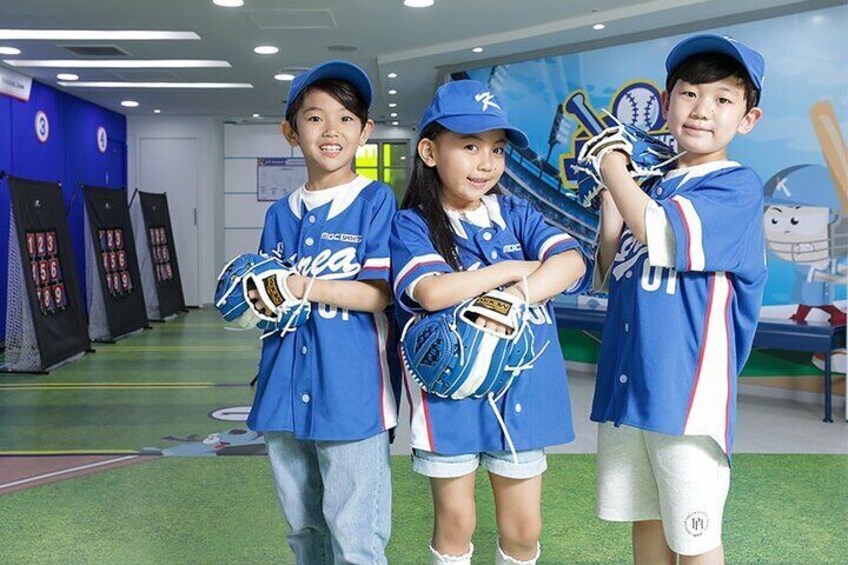 Three children in blue baseball uniforms standing in a sports facility with green flooring and walls decorated with sports-themed murals. 5