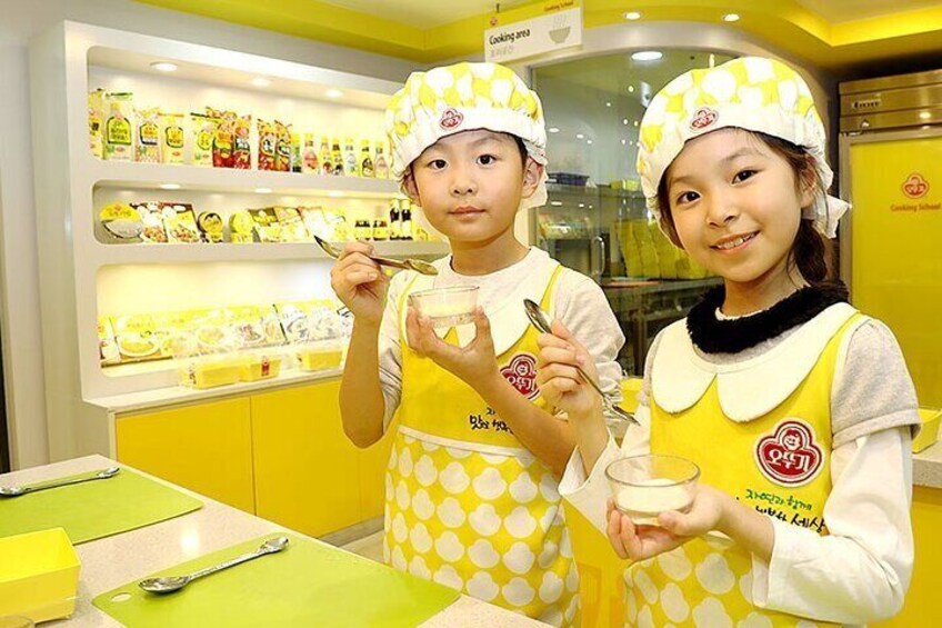 Two people in yellow aprons and chef hats in a kitchen area with shelves of food products. 1