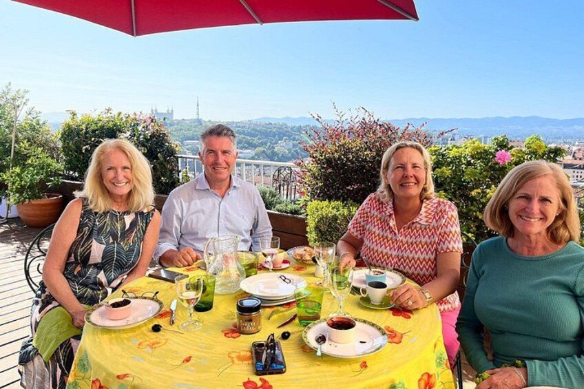 Four people dining at an outdoor table with a yellow tablecloth, surrounded by potted plants and a scenic view. 2