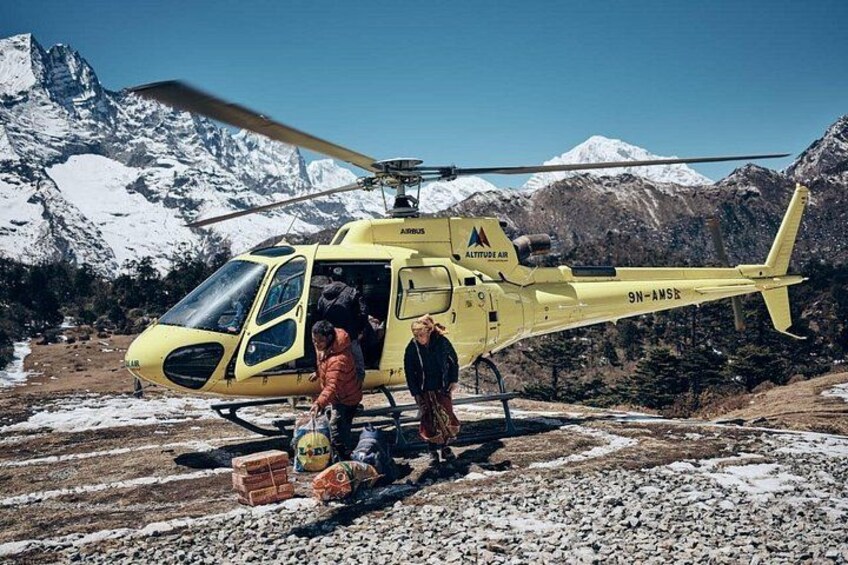 A yellow helicopter with the text 'ALITUDES AIR' and '9N-AMS' on its side, parked on a snowy ground with mountains in the background. 1