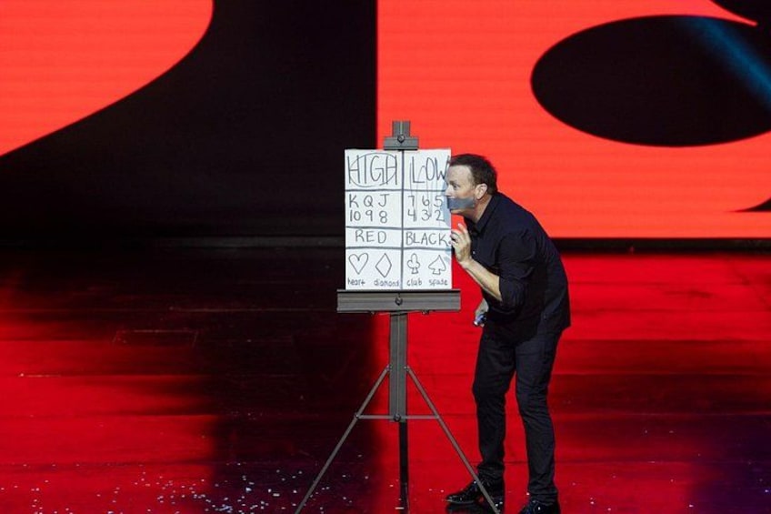 Een persoon staat op een podium, met een microfoon, een bord met tekst en symbolen, en een rode achtergrond. 3