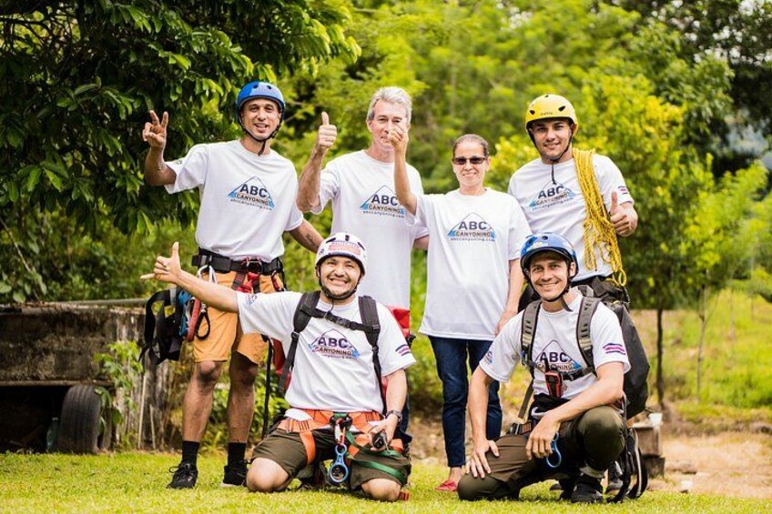 A group of people in outdoor gear, some wearing helmets, standing and sitting on grass with trees in the background. 5