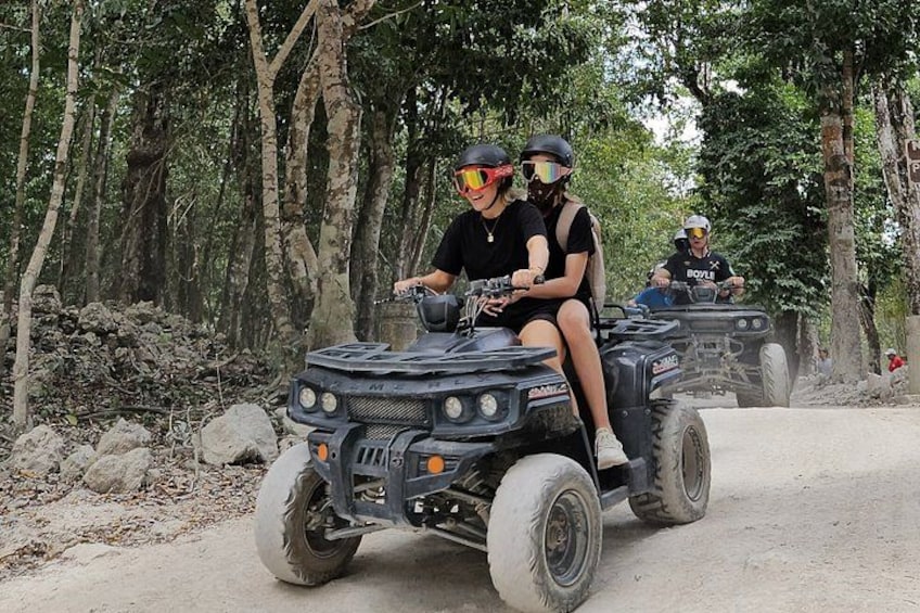 Two people riding ATVs on a dirt road surrounded by trees. 1