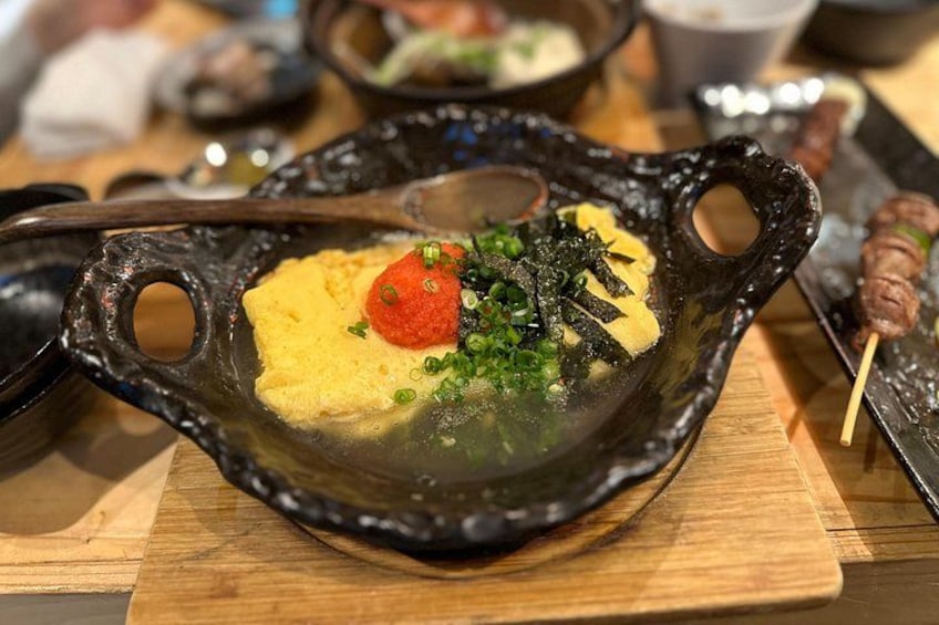 Un plat en céramique noire contenant une substance jaune, garni d’une décoration rouge et d’oignons verts, le tout posé sur une table en bois. 4