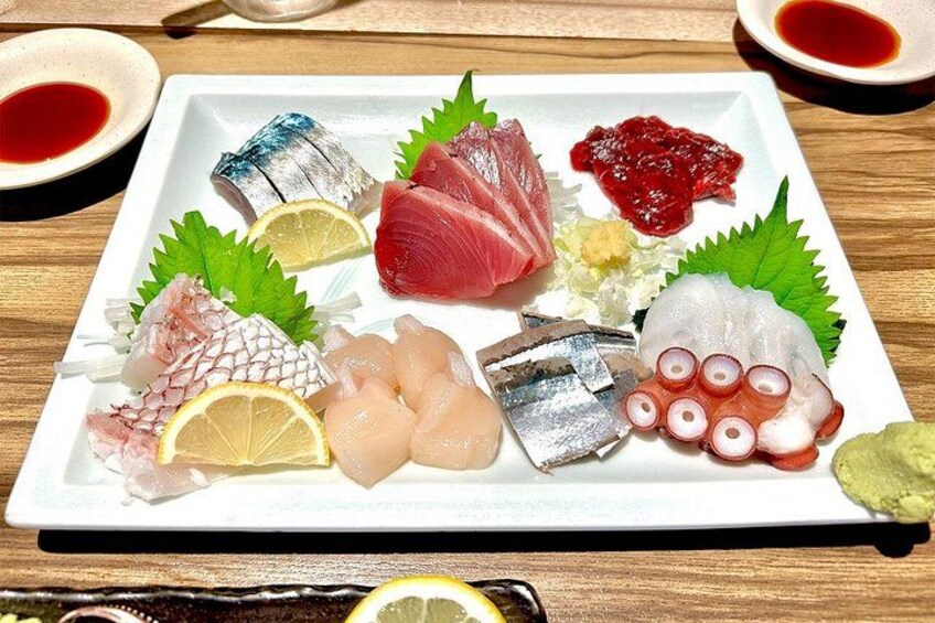 Une variété de sashimis présentés sur une assiette blanche, accompagnés de feuilles vertes, de tranches de citron et de petits ramequins de sauce. 2