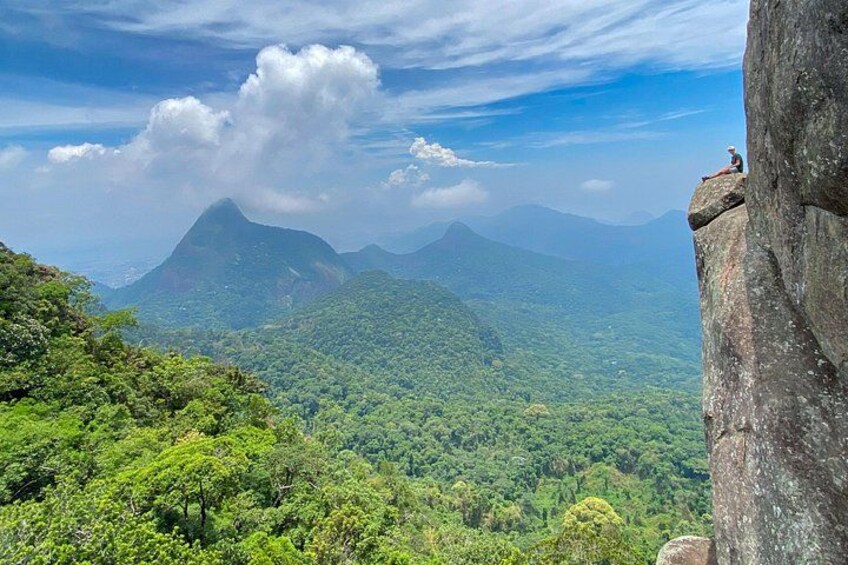 A person sitting on a rock ledge overlooking a dense forest and distant mountains. 5