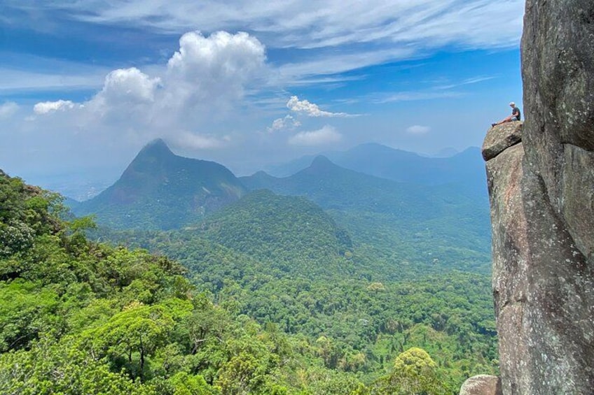 A person sitting on a rock ledge overlooking a dense forest and distant mountains under a partly cloudy sky. 4