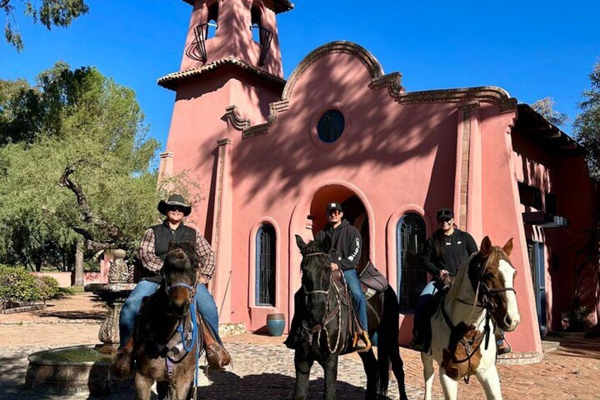 A pink church with a bell tower, people on horseback, and a clear blue sky. 2
