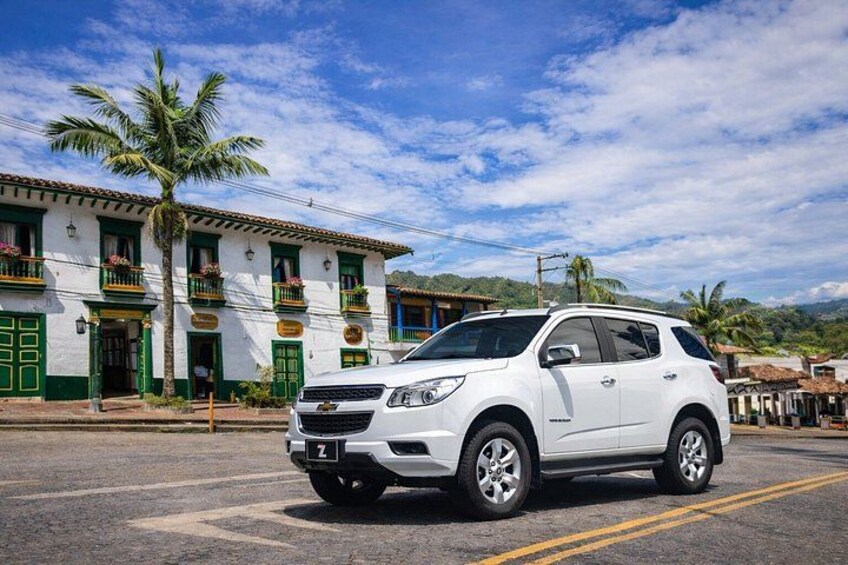 A white SUV parked on a street with a building featuring green doors and balconies in the background. 1