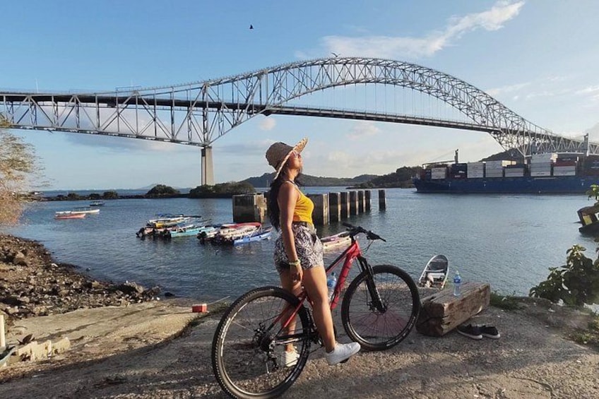 Une personne se tient à côté d’un vélo, près d’une rivière, avec un pont en arrière-plan. 1