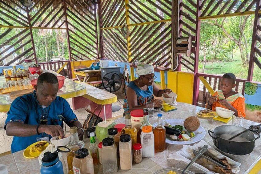 Personas preparando comida en un entorno rústico al aire libre, con techo de paja y construcciones de madera. 4
