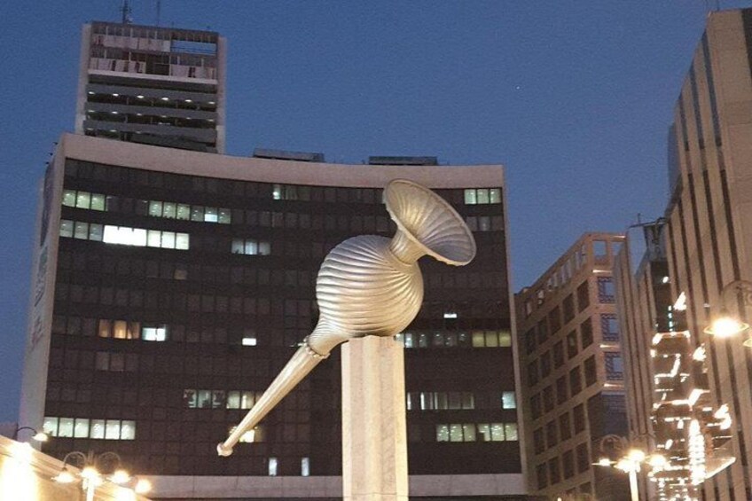 En skulptur som föreställer en fågel med lång hals och stort huvud, placerad framför en bakgrund av moderna byggnader i skymningen. 2