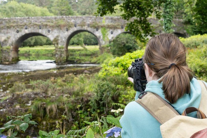 A person taking a photograph with a camera, a stone bridge, and greenery in the background. 5