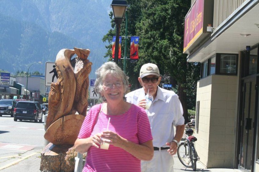 Two people standing next to a large wooden sculpture on a street with a mountain in the background. 2