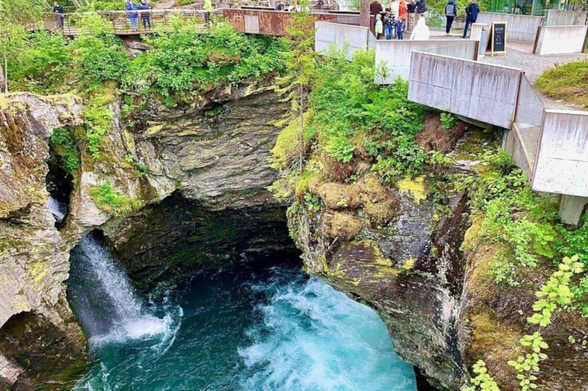 Ein Wasserfall stürzt über eine felsige Klippe, umgeben von üppiger Vegetation und einer Aussichtsplattform mit Personen. 2