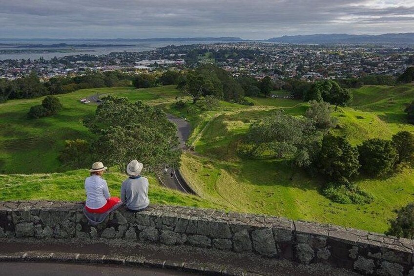 Two people sitting on a stone wall overlooking a green landscape with a river and a town in the distance. 1