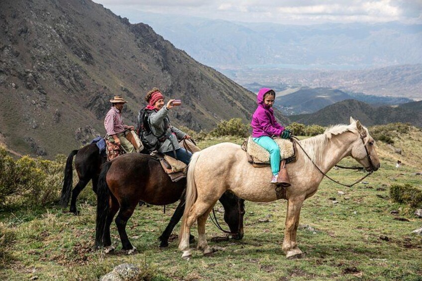 Three people riding horses on a grassy hillside with mountains in the background. 4