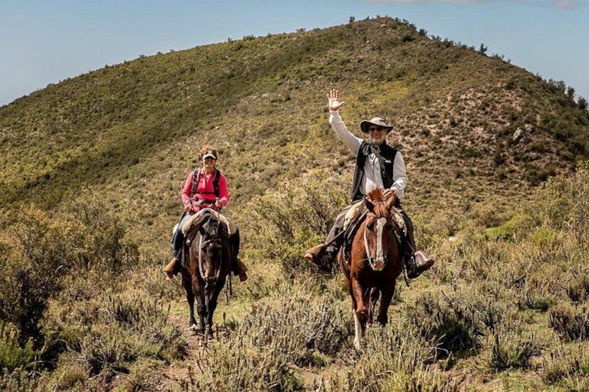 Two people horseback riding on a grassy hill with sparse vegetation. 1